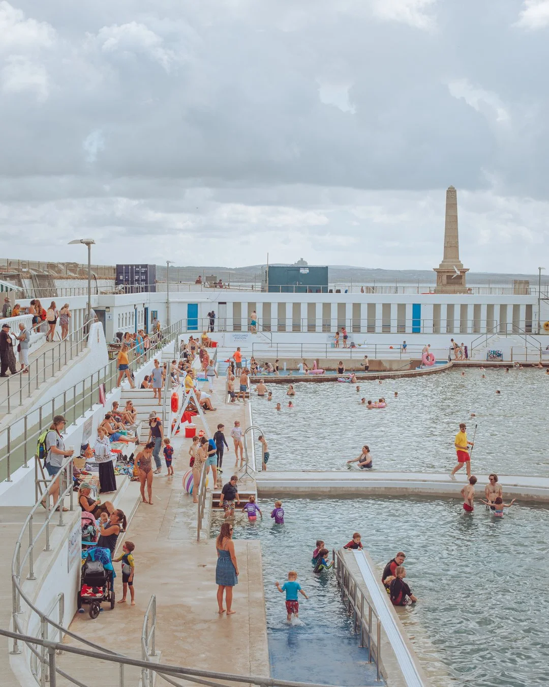 People enjoy swimming and relaxing at a seaside outdoor pool area with cloudy skies.