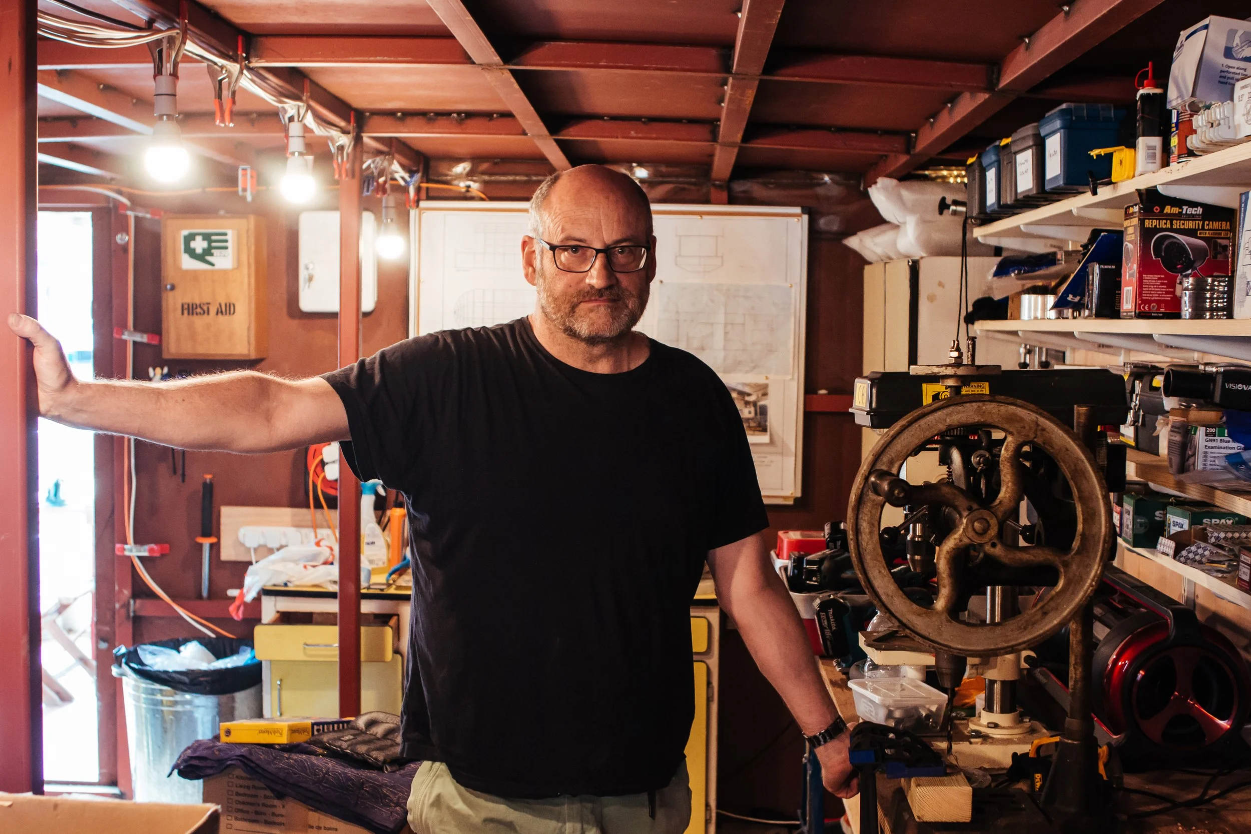 A man with glasses and a beard standing in a cluttered workshop or garage, holding onto a workbench with one hand and touching a large woodworking or metalworking machine with the other.