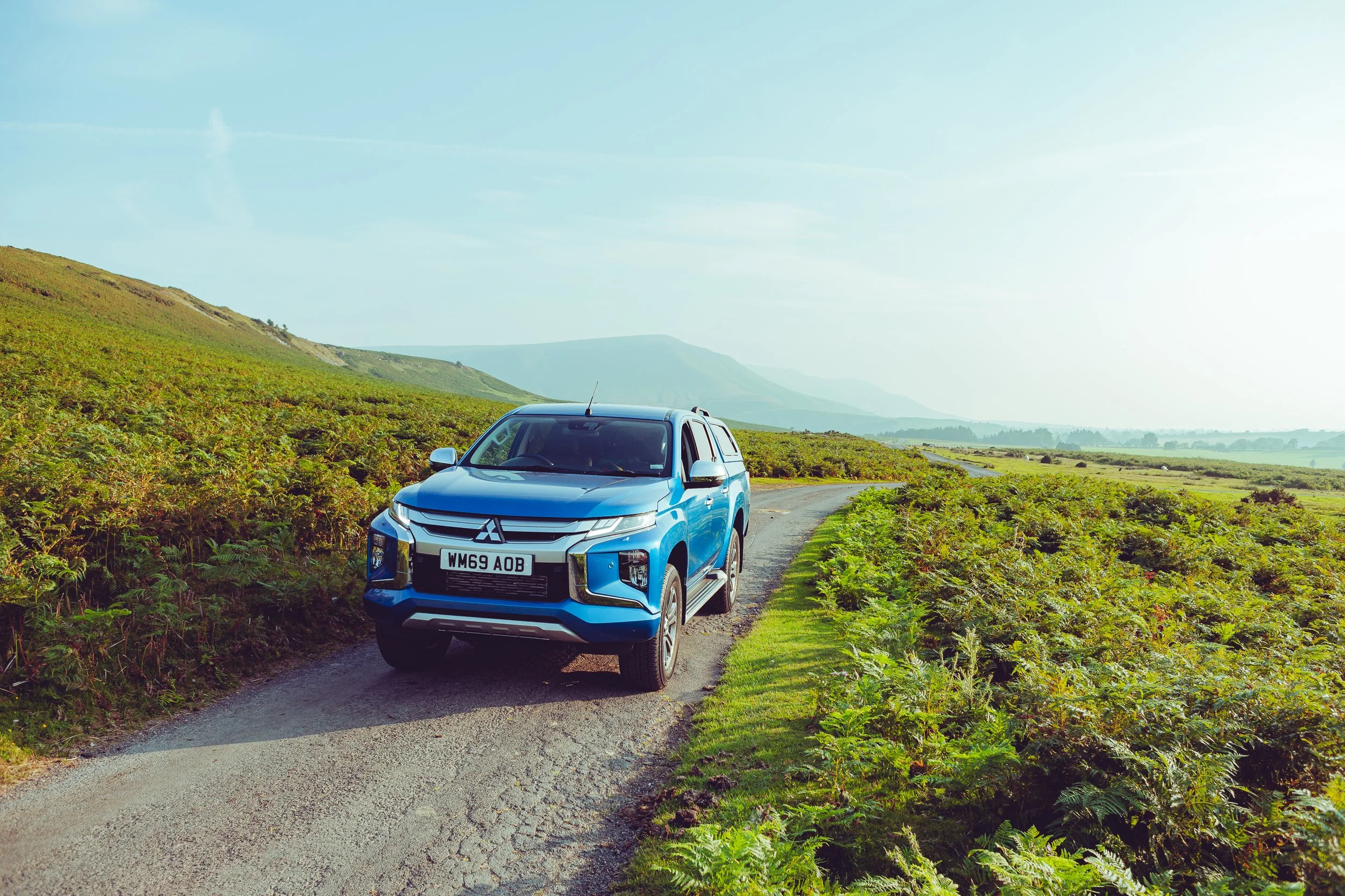 A blue Mitsubishi SUV on a narrow country road surrounded by green fields and rolling hills in the distance under a partly cloudy sky.