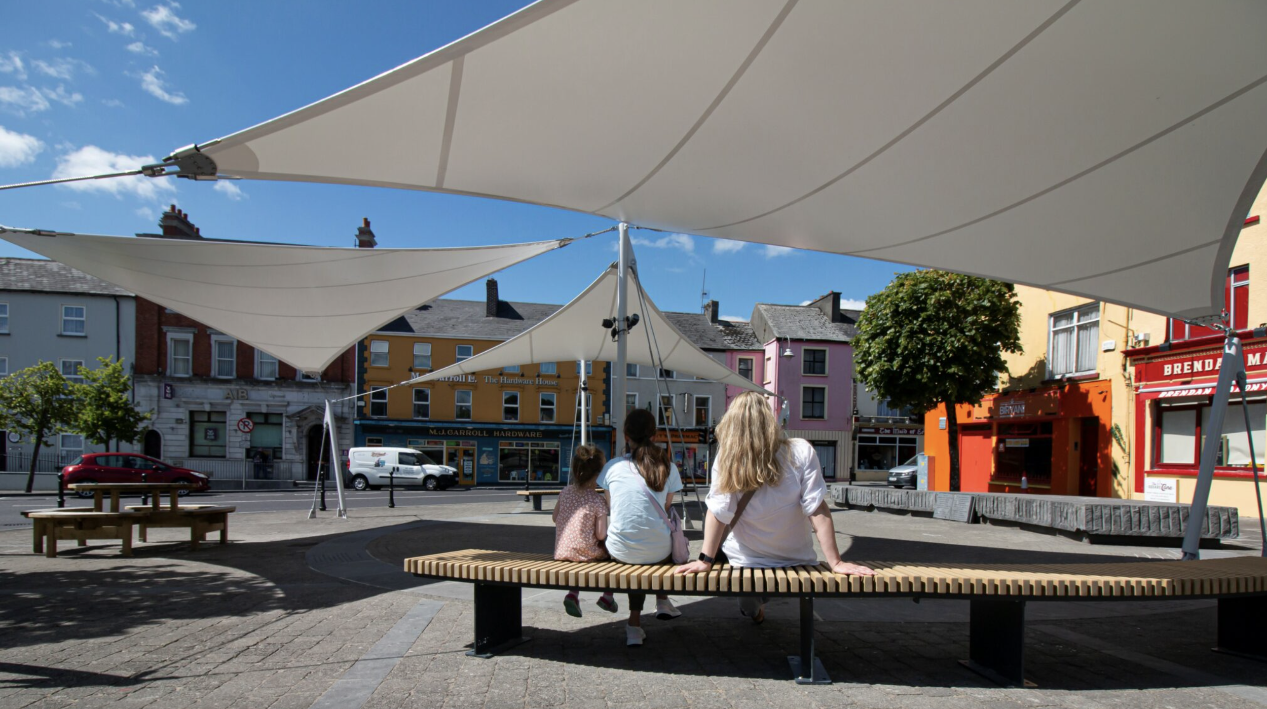 A group of three people, two women and a girl, sitting on a curved park bench under large white shade sails in a colorful urban plaza with storefronts and cars in the background.
