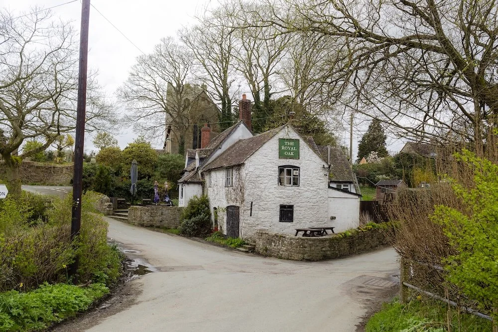 Small white house called "The Royal Oak" with a green sign, surrounded by trees and greenery, at a fork in a rural road.