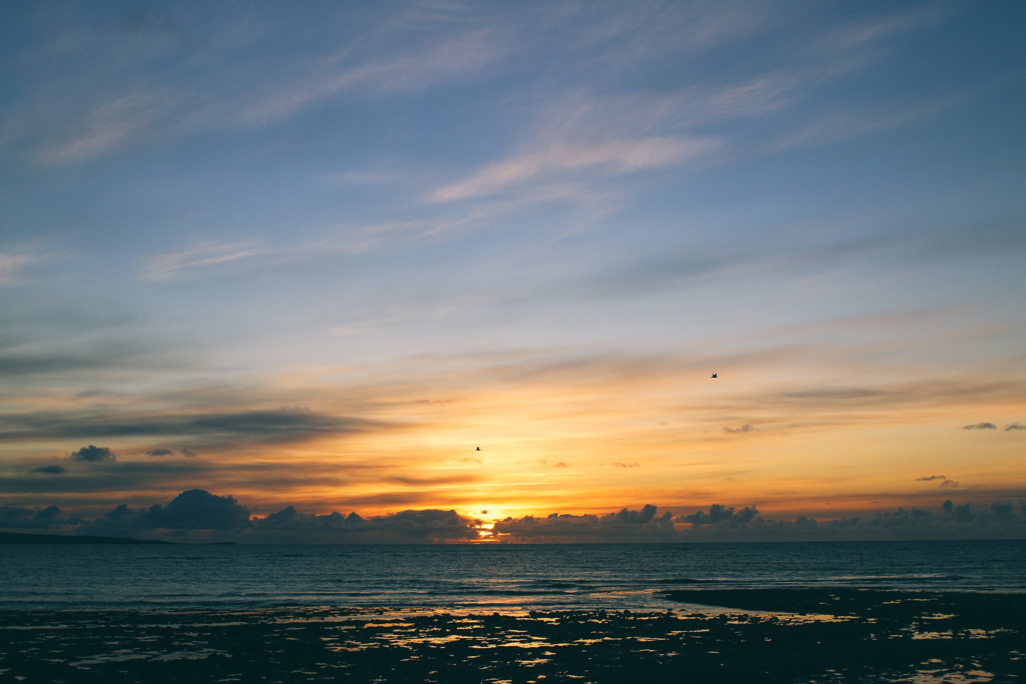 Sunset over the ocean with a partly cloudy sky, two birds flying, and reflections on the water.