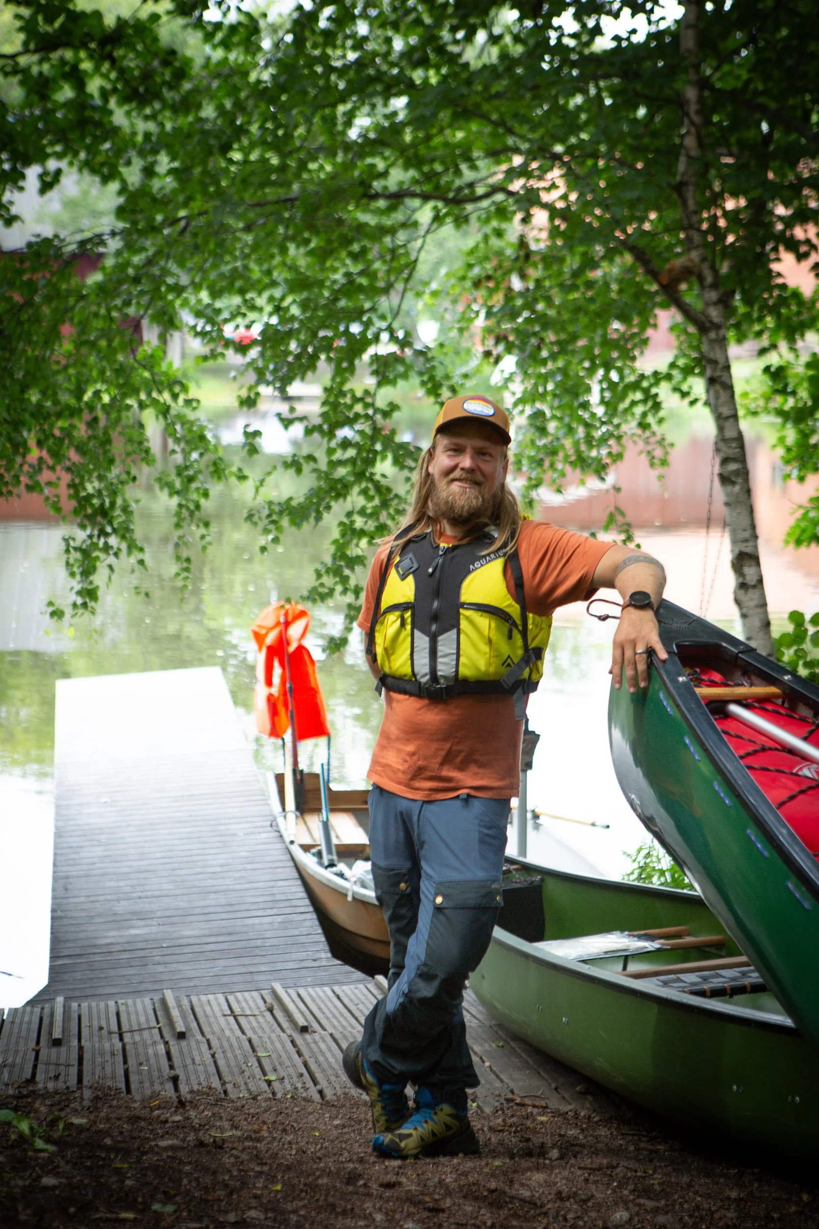 A man standing next to two kayaks on a dock near a body of water, under green leafy trees, smiling and wearing outdoor gear.