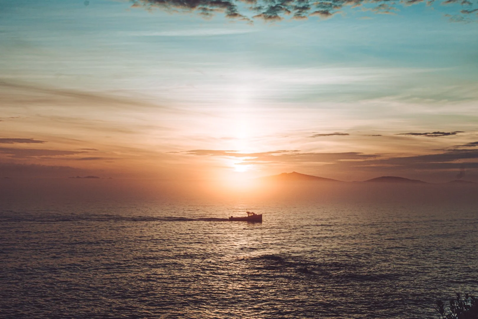 A boat sailing on the ocean during sunset with hills in the background and colorful sky.