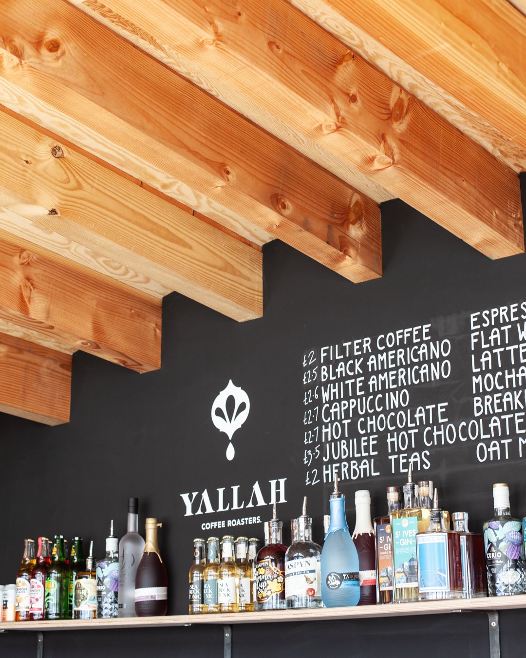 A black wall with a menu written in white chalk, showing different coffee and tea options and prices. Displayed on the wall is a white logo and hexagon symbol for YALLAH Coffee Roasters. Several bottles and containers are on a shelf below.
