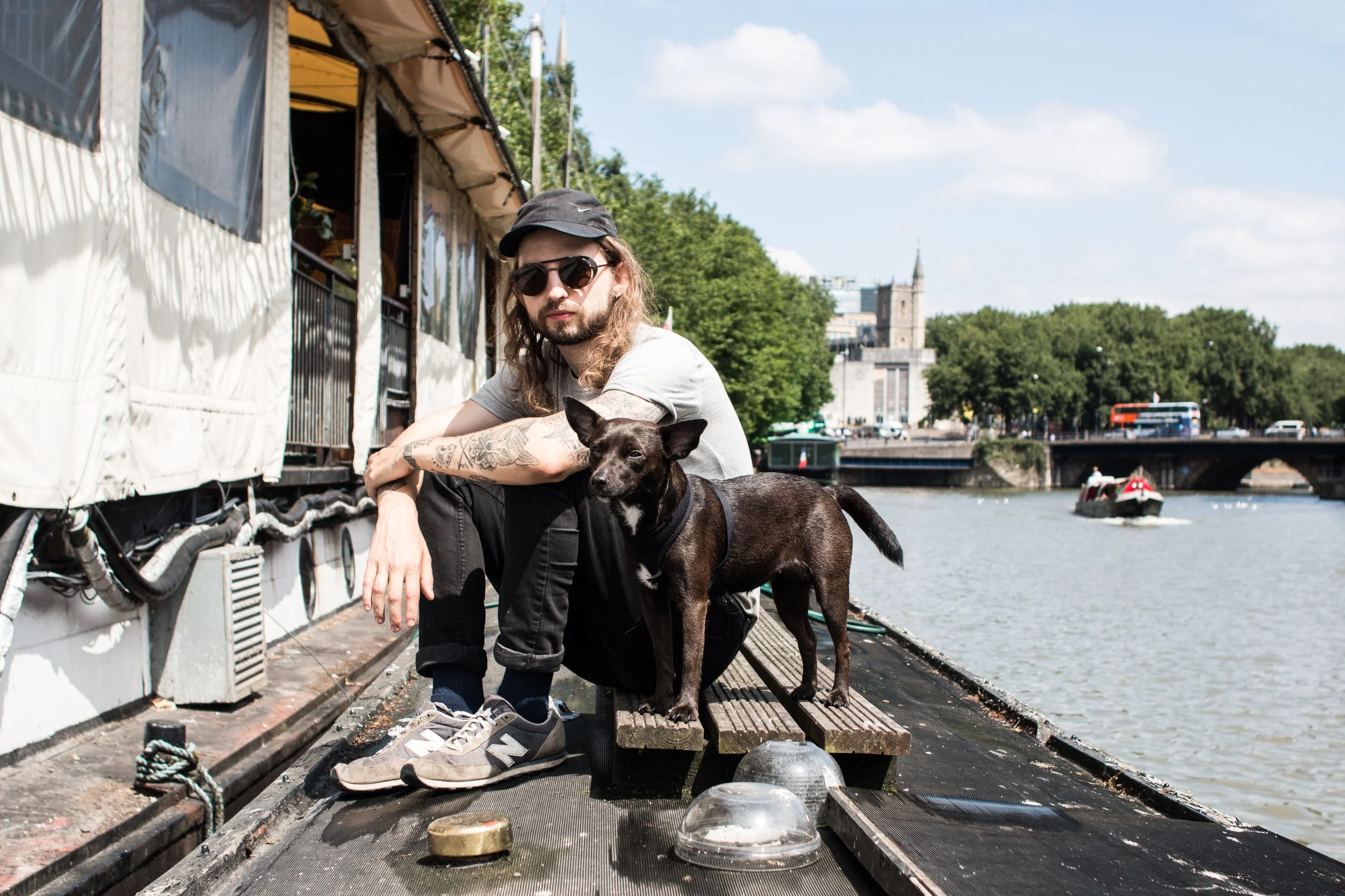 A man with tattoos and long hair, wearing sunglasses, a gray T-shirt, black pants, and sneakers, sitting on a boat with his dog by a river in an urban area. The man is wearing a black cap, and the dog is a small, dark-colored, mixed breed standing be