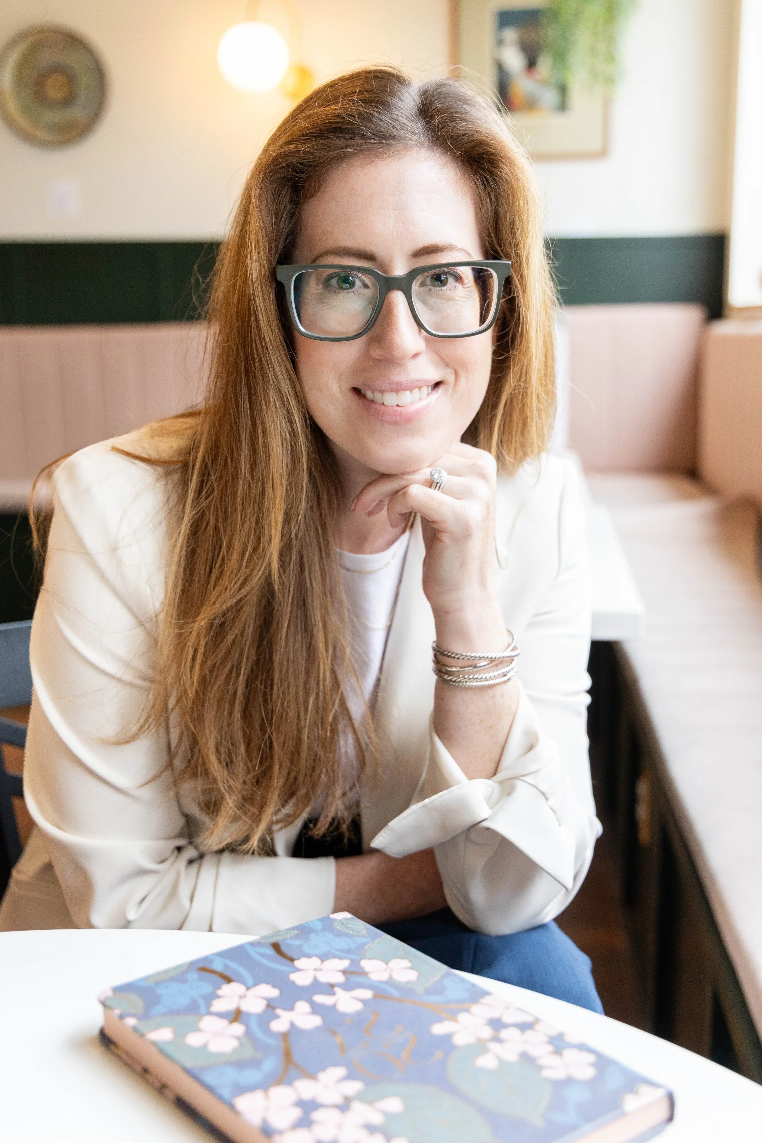 A woman with long red hair and glasses sitting at a table in a cozy cafe, resting her chin on her hand, with a floral notebook on the table in front of her.
