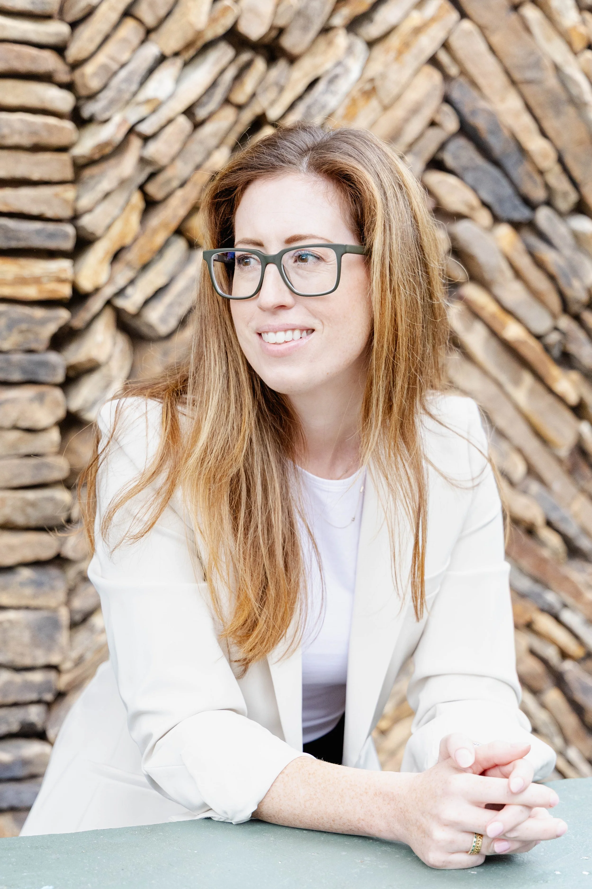A woman with long red hair and glasses wearing a white blazer sitting at a table in front of a stacked wood wall.
