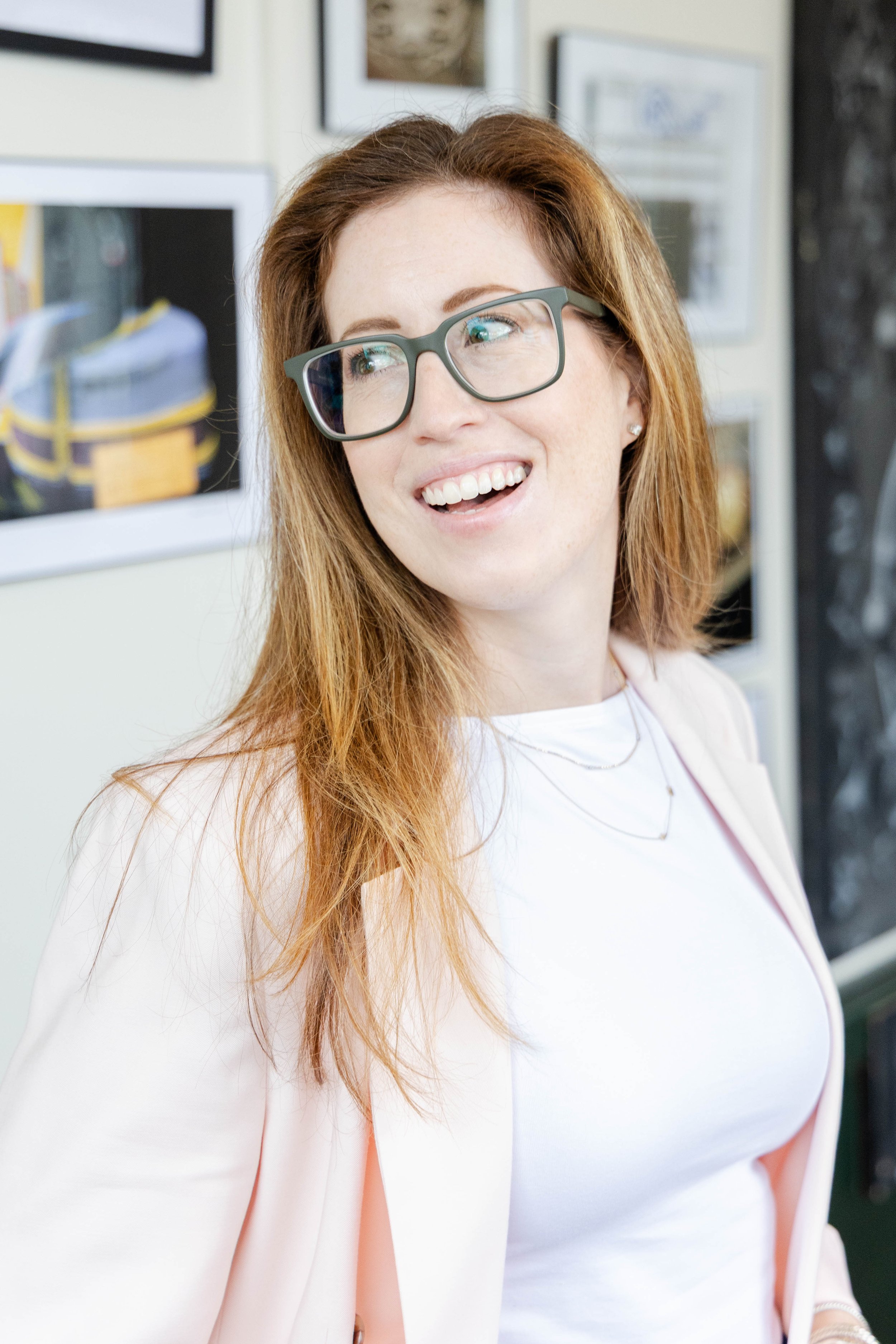 A woman with long red hair wearing glasses, smiling, and dressed in a white top and light pink blazer.