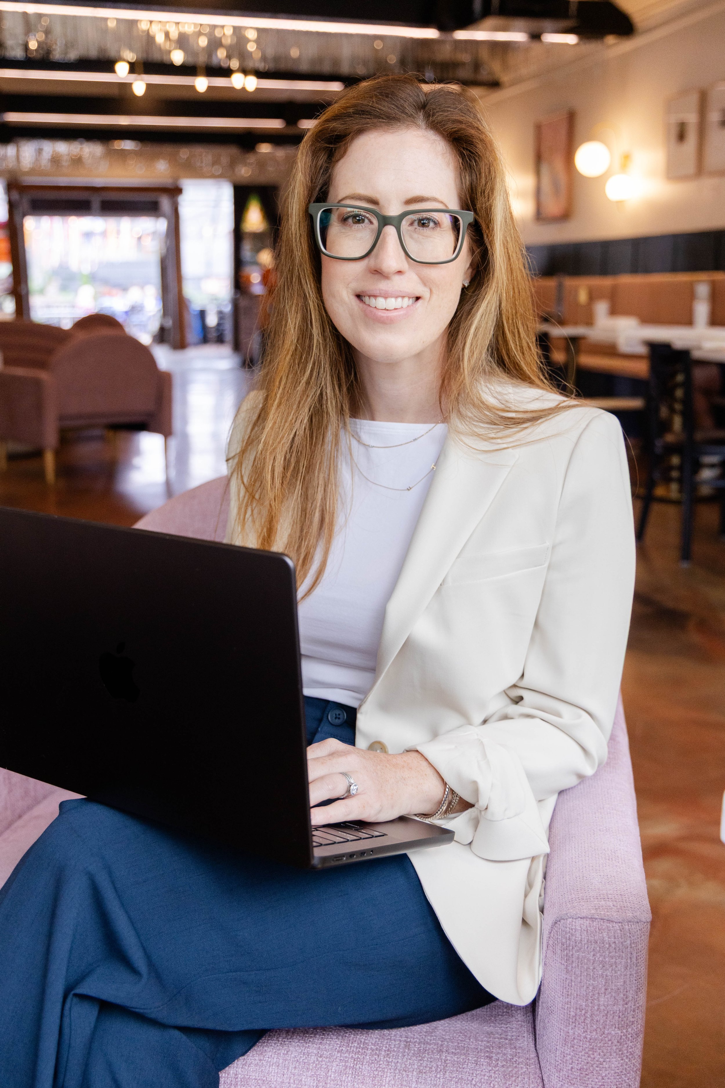 A woman with long red hair and glasses sitting in a cafe, smiling at the camera, with a laptop on her lap.