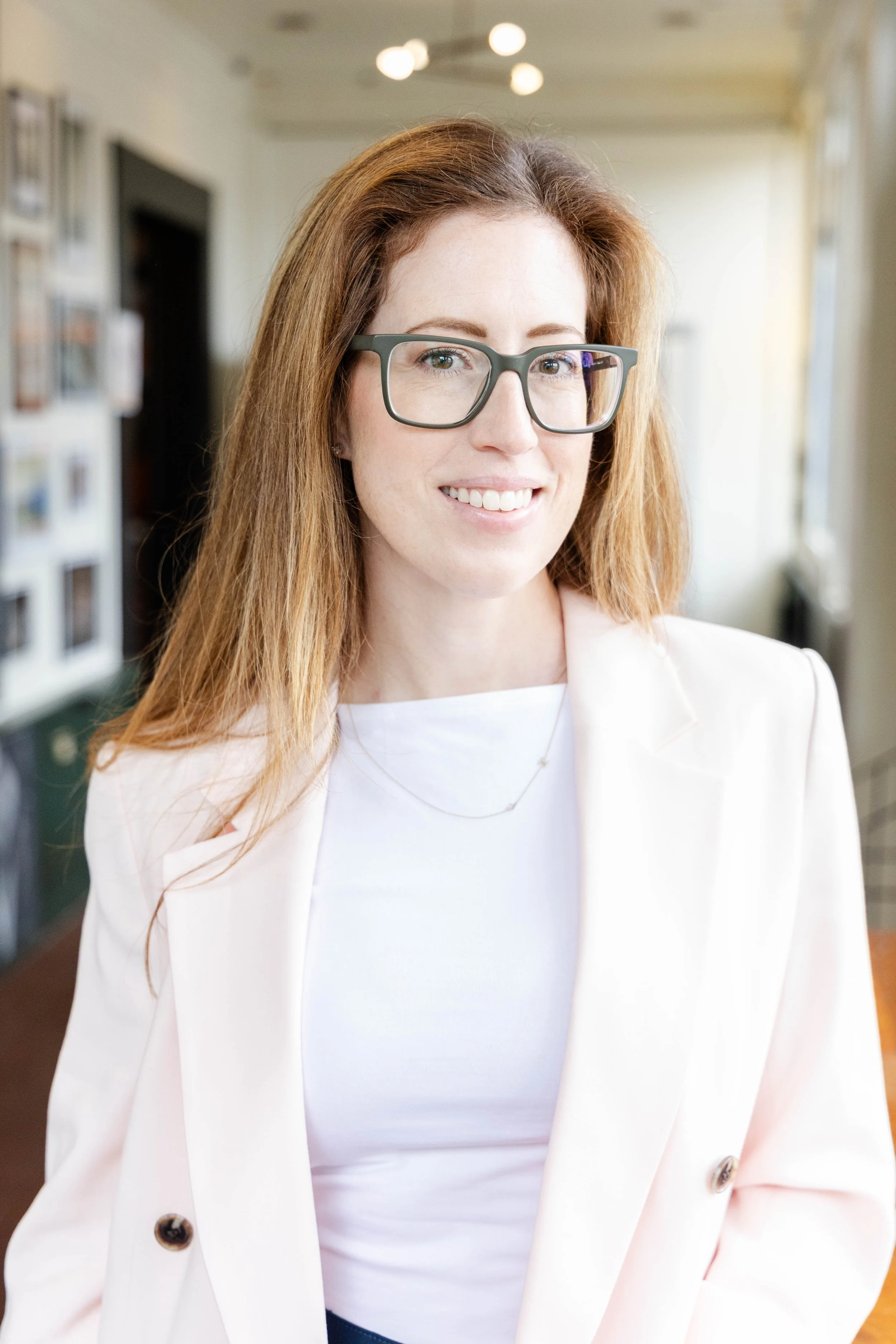 A woman with red hair, glasses, and a white blazer smiling indoors with a blurred background of photo frames and a window.