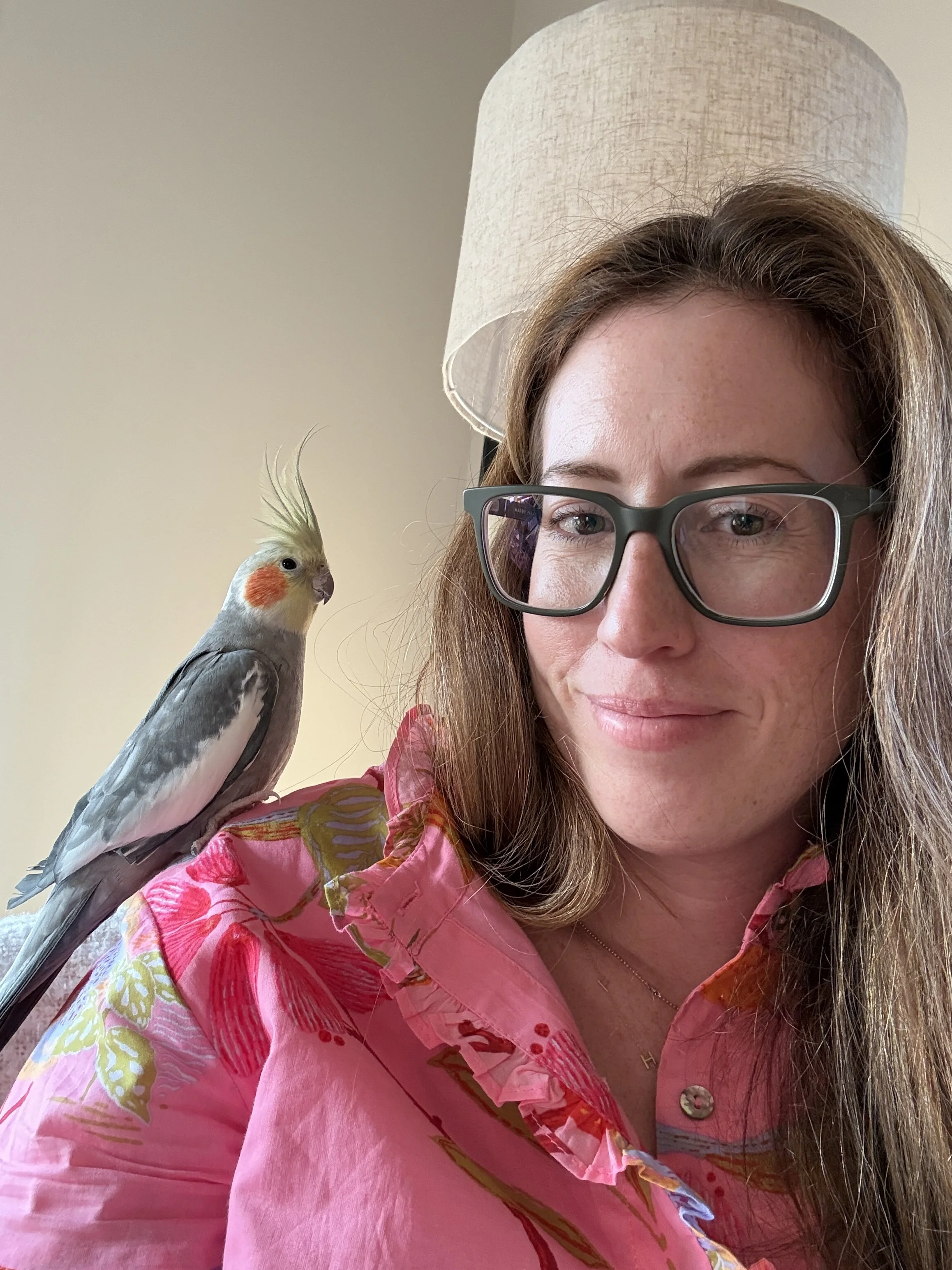 A woman with long brown hair, wearing glasses and a pink floral shirt, is smiling slightly. A cockatiel bird with yellow crest, orange cheeks, and gray and white feathers is perched on her shoulder, looking slightly to the side.