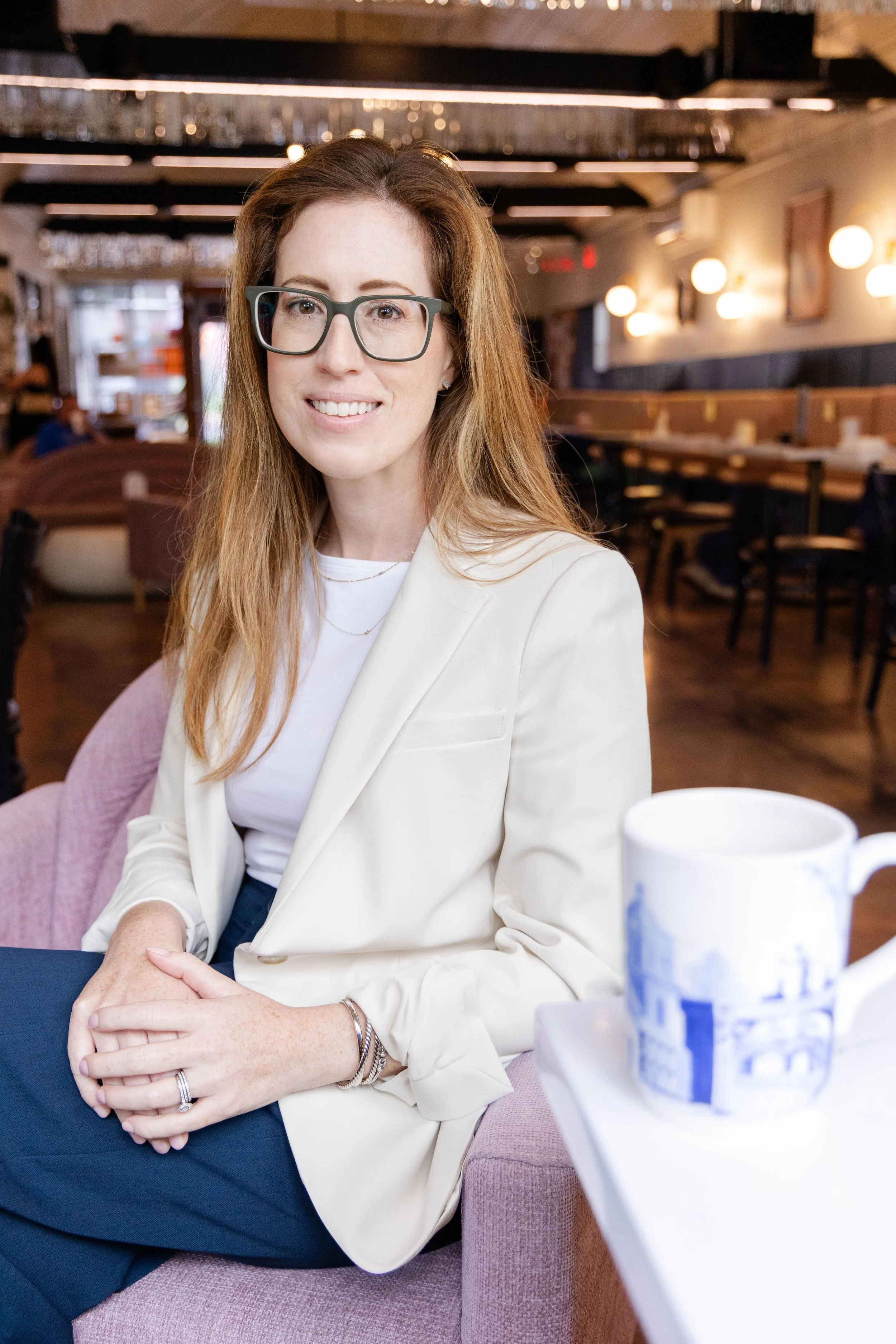 A woman with red hair, glasses, and fair skin smiling while sitting in a restaurant. She is wearing a white blazer and has rings and bracelets on her hands.