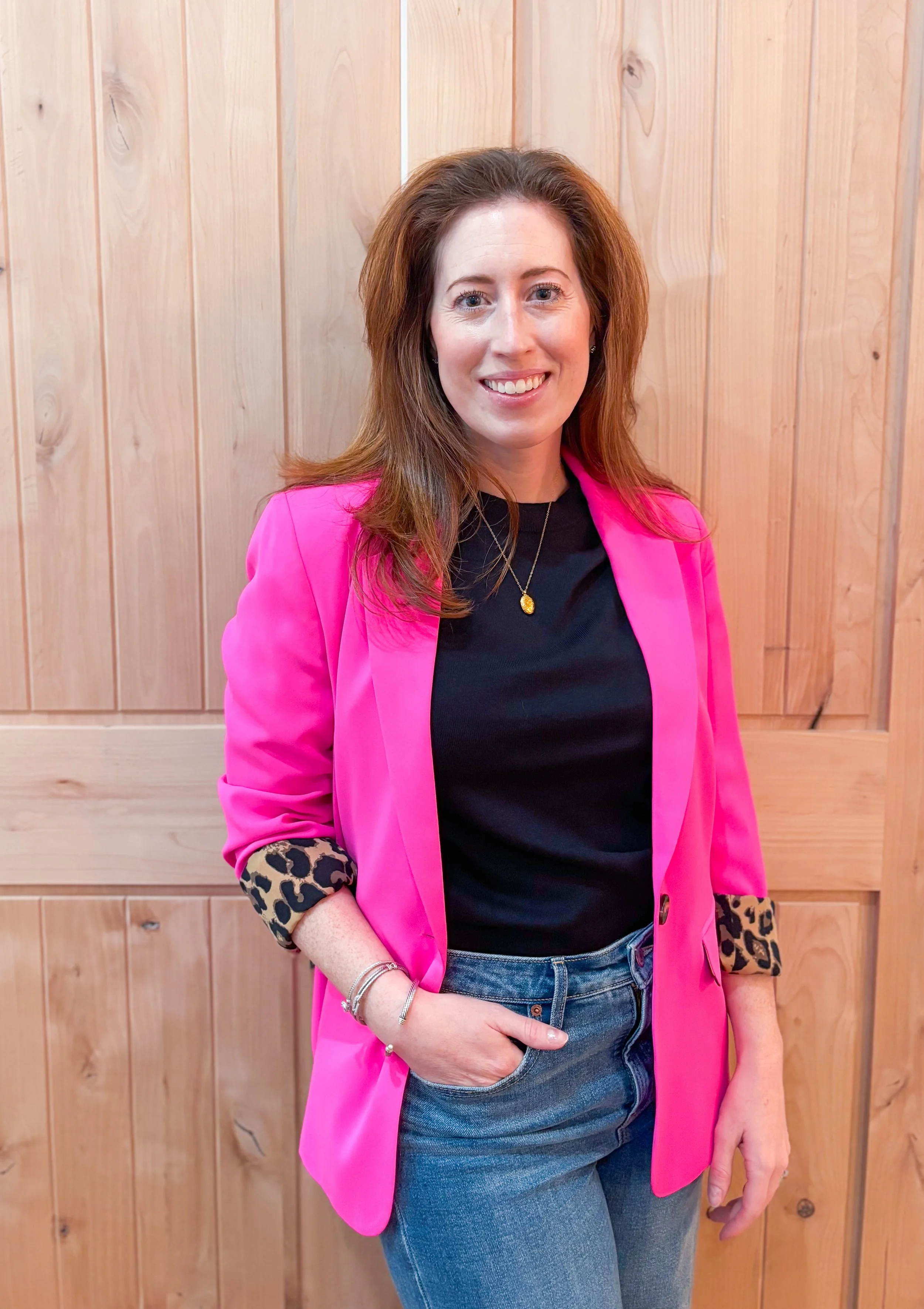 A woman with red hair wearing a bright pink blazer with leopard print sleeves, a black top, and blue jeans standing in front of a wooden wall, smiling at the camera.