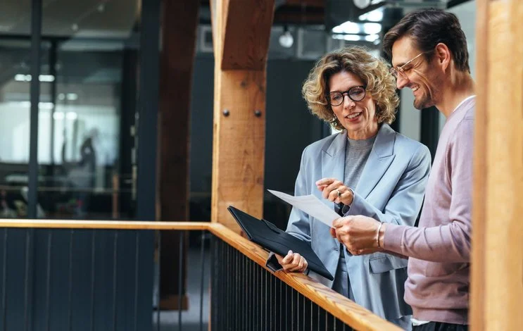 Two colleagues, a woman with curly gray hair and glasses wearing a light blue blazer, and a man with dark hair and glasses wearing a light purple sweater, are both smiling and looking at a document together in an office setting with wooden beams and glass windows.