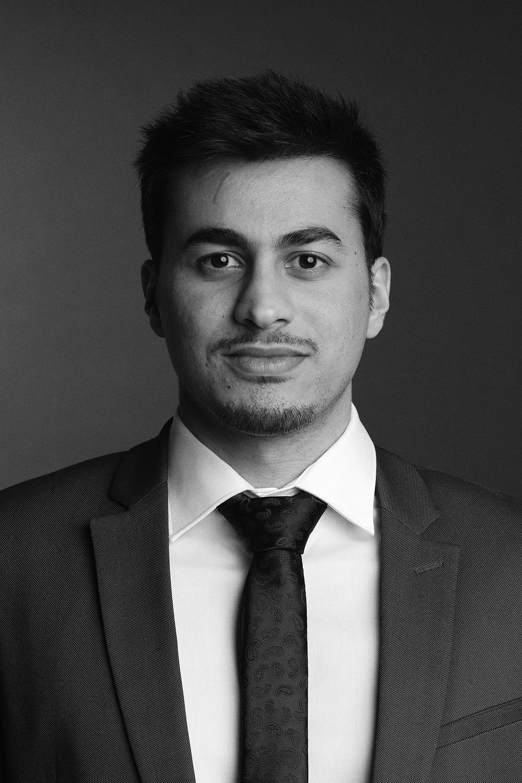 Black and white portrait of a young man in a suit, white shirt, and tie, looking directly at the camera with a neutral expression.
