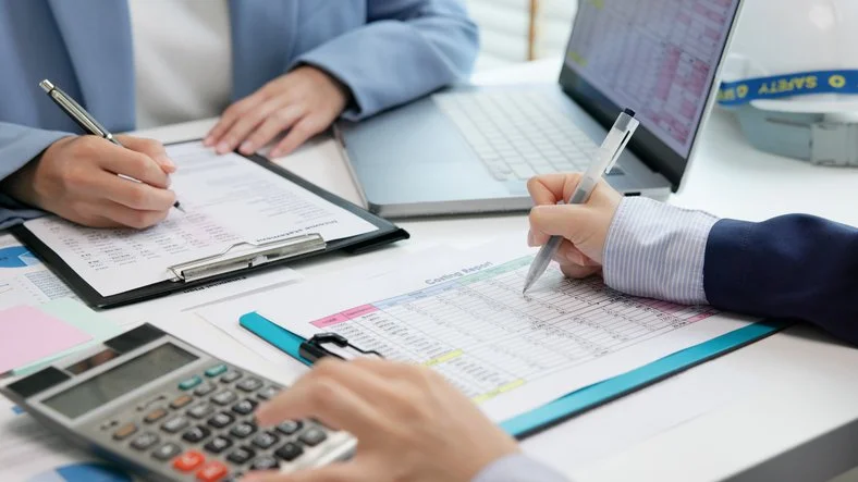People working on financial documents with a calculator, a pen, and a laptop on a desk.