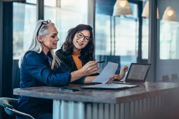 Two women collaborating at a desk with a laptop and papers in a modern office.