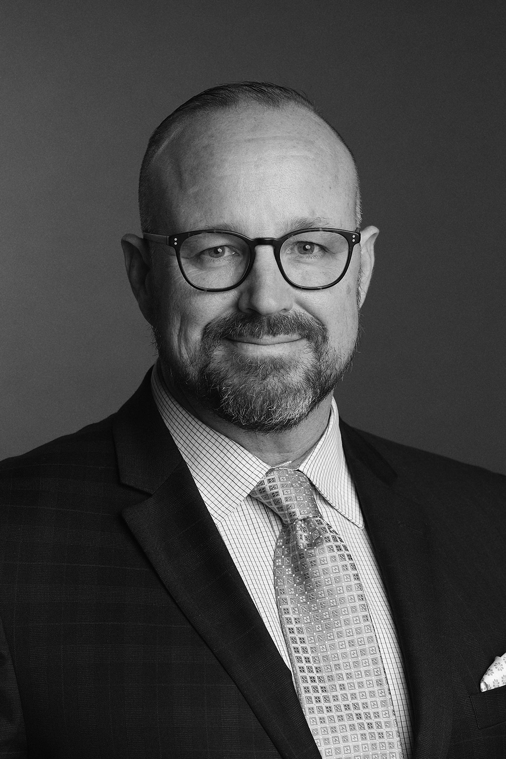 Black and white professional headshot of a middle-aged man with glasses, a beard, and a mustache, wearing a suit, dress shirt, and patterned tie.