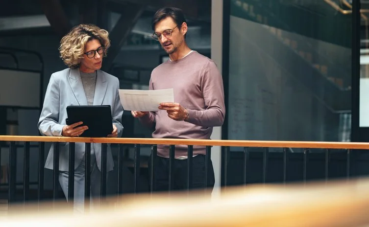 Two people, a woman in a light blue suit and a man in glasses and a pink sweater, are standing on a balcony looking at documents.