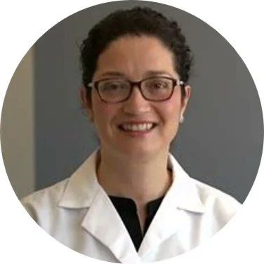 A woman with curly dark hair wearing glasses and a white lab coat, smiling at the camera.