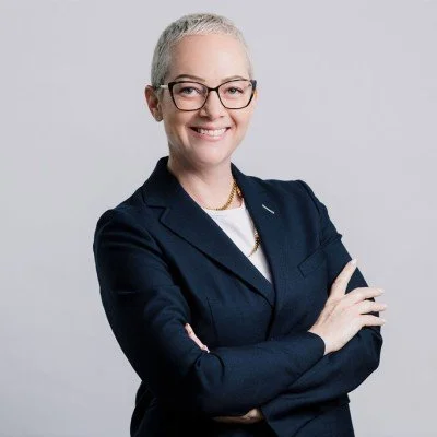 Portrait of a smiling woman with short gray hair, wearing glasses and a navy blazer, crossing her arms against a light gray background.