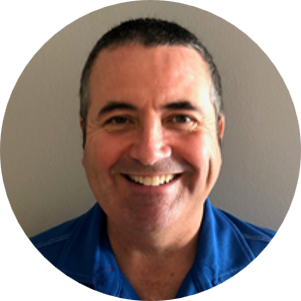 Portrait of a smiling man with short dark hair, wearing a blue shirt, standing against a gray background.