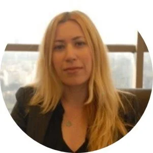 A woman with long blonde hair sitting indoors near a window with a blurred city background.