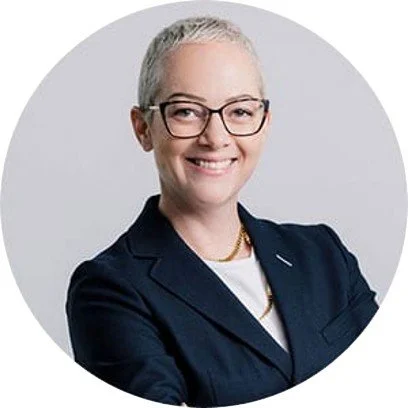 Professional woman with short gray hair and glasses, smiling in a navy blazer against a light background.