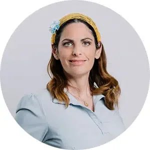 A woman with shoulder-length brown hair wearing a blue headband with a flower, a light blue blouse, and a subtle smile, posed against a light gray background.