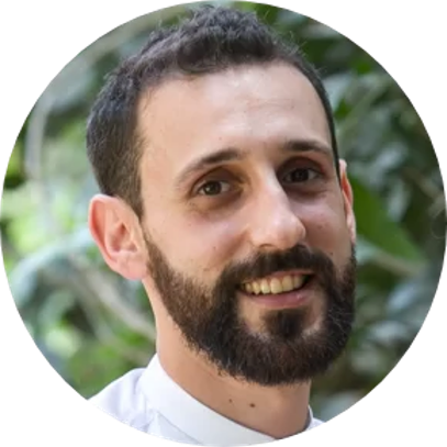 A smiling man with dark hair and a beard outdoors, with greenery in the background.