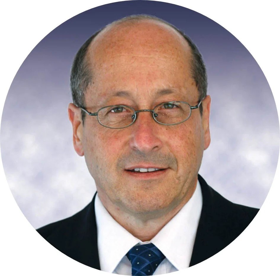 A middle-aged man with glasses, wearing a suit, white shirt, and a blue tie, is smiling in front of a cloudy sky background.