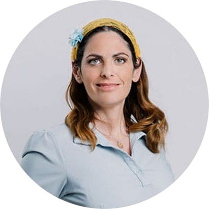 Woman with brown hair, wearing a light blue blouse and a yellow headband with a blue flower, posing against a gray background.