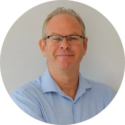 A middle-aged man with glasses, short gray hair, and a light blue button-down shirt smiling with arms crossed, standing against a plain light background.