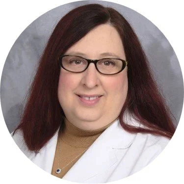 A woman with long red hair and glasses, wearing a white coat and a beige top, smiling at the camera.