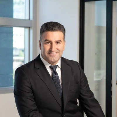 Professional man in a black suit and tie smiling in an office setting with large windows