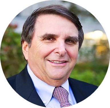 A smiling middle-aged man in a suit and tie outdoors with greenery in the background.