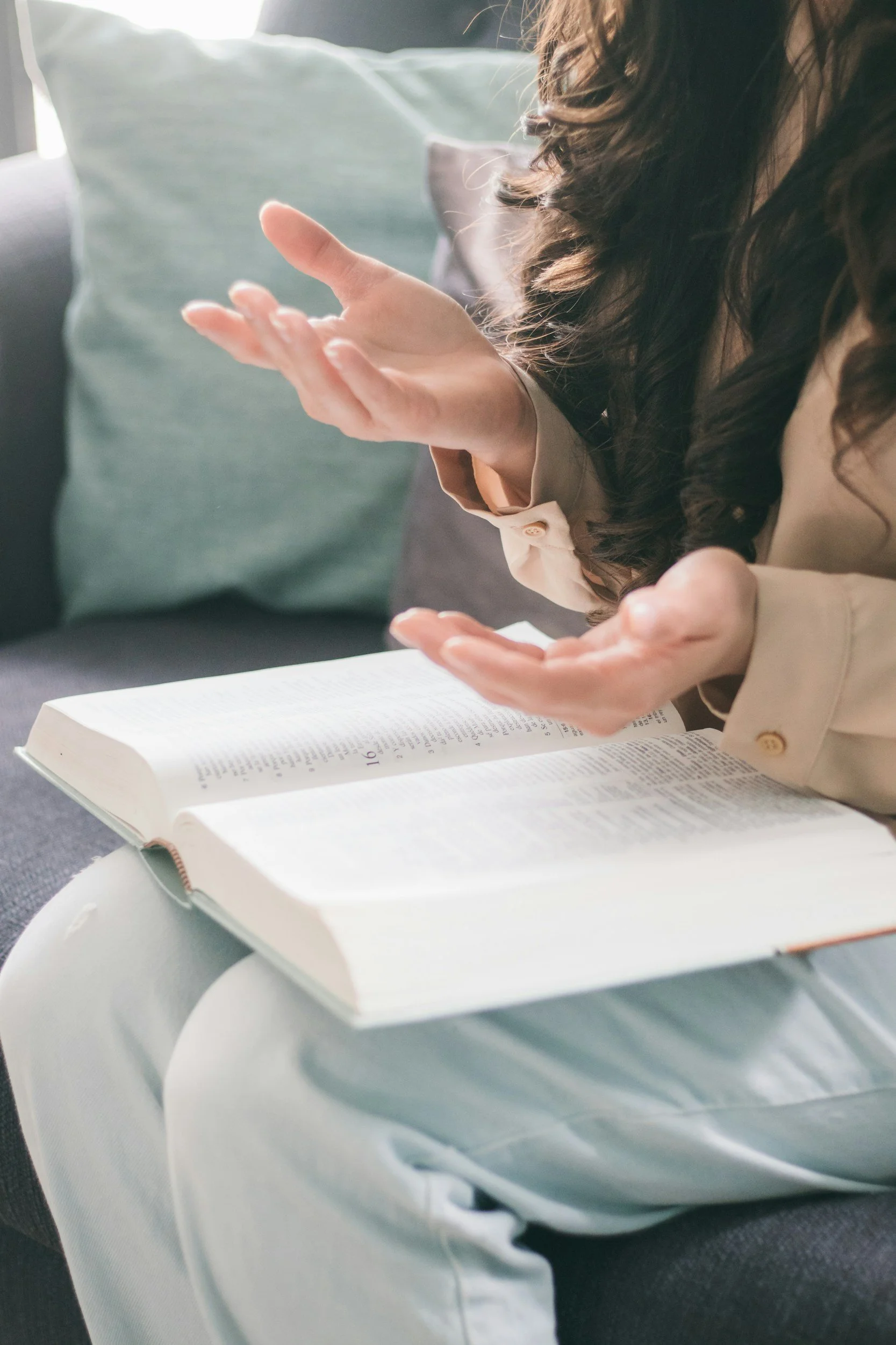A woman with long, curly brown hair reading an open book, sitting on a gray couch with light green and gray pillows visible in the background.