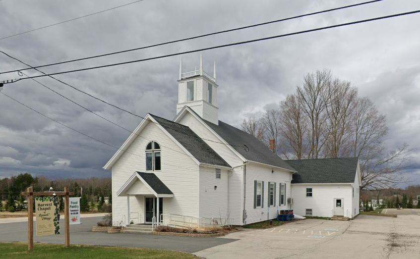 White church with steeple and other buildings, parking lot in front, cloudy sky, leafless trees in the background.