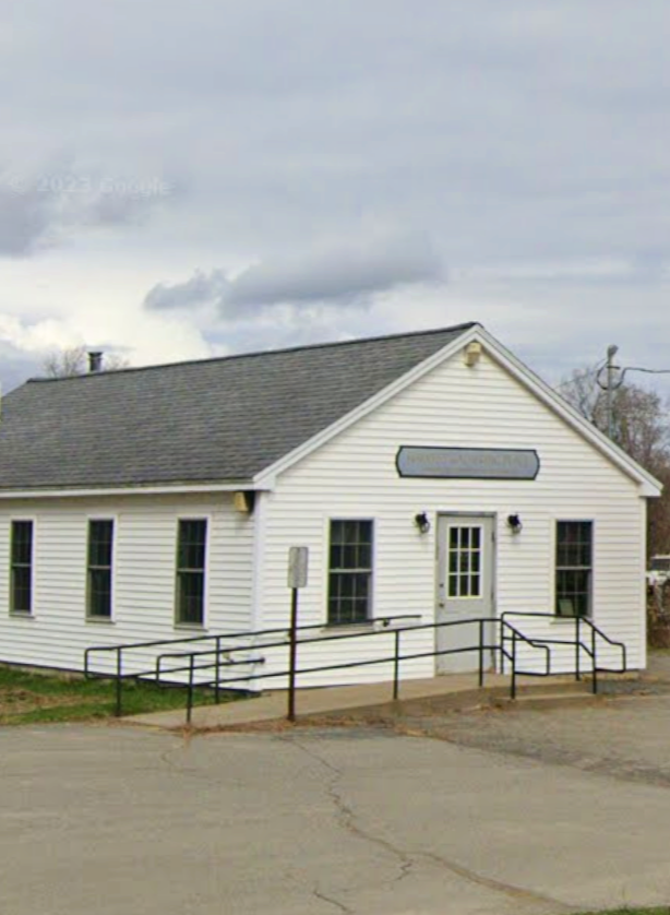 Small white building with a gray roof, three windows on each side, a door with a small window, black railings leading to the entrance, and an empty sign above the door, under a cloudy sky.