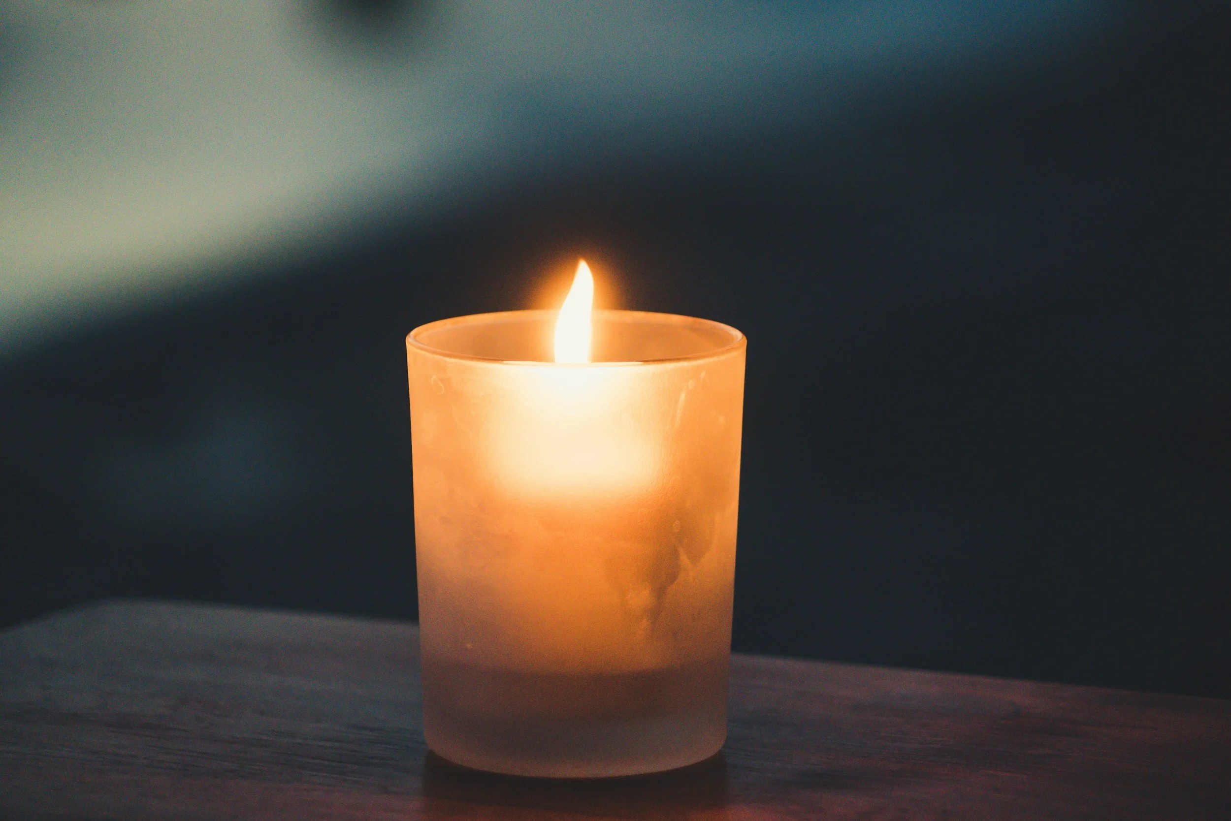 A lit candle in a frosted glass holder on a wooden surface with a dark background.