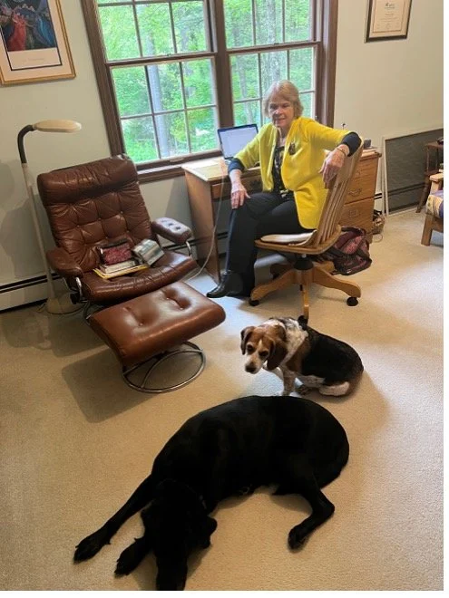 An elderly woman in a yellow jacket sitting at a desk by a window with two dogs lying on the carpet in front of her, a black Labrador and a small dog with black, white, and brown fur.