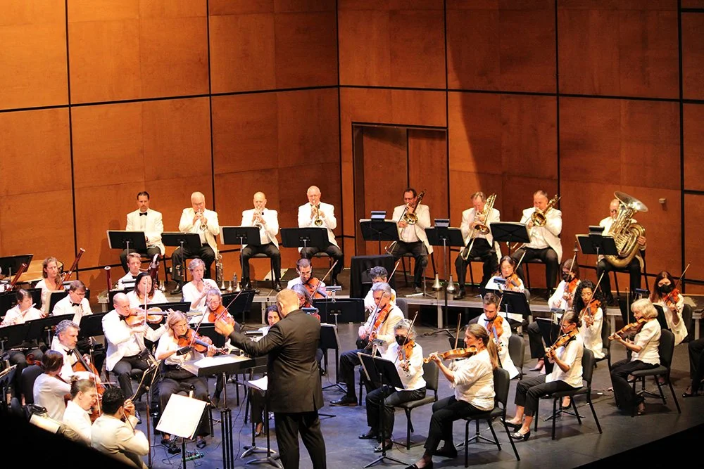 An orchestra performing on stage in a concert hall, with musicians playing string, brass, and woodwind instruments, and a conductor leading them.