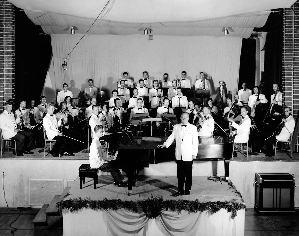 Black and white photo of an orchestra on stage with a conductor standing in front of a grand piano. The orchestra members wear formal attire, and the stage is decorated with garland.