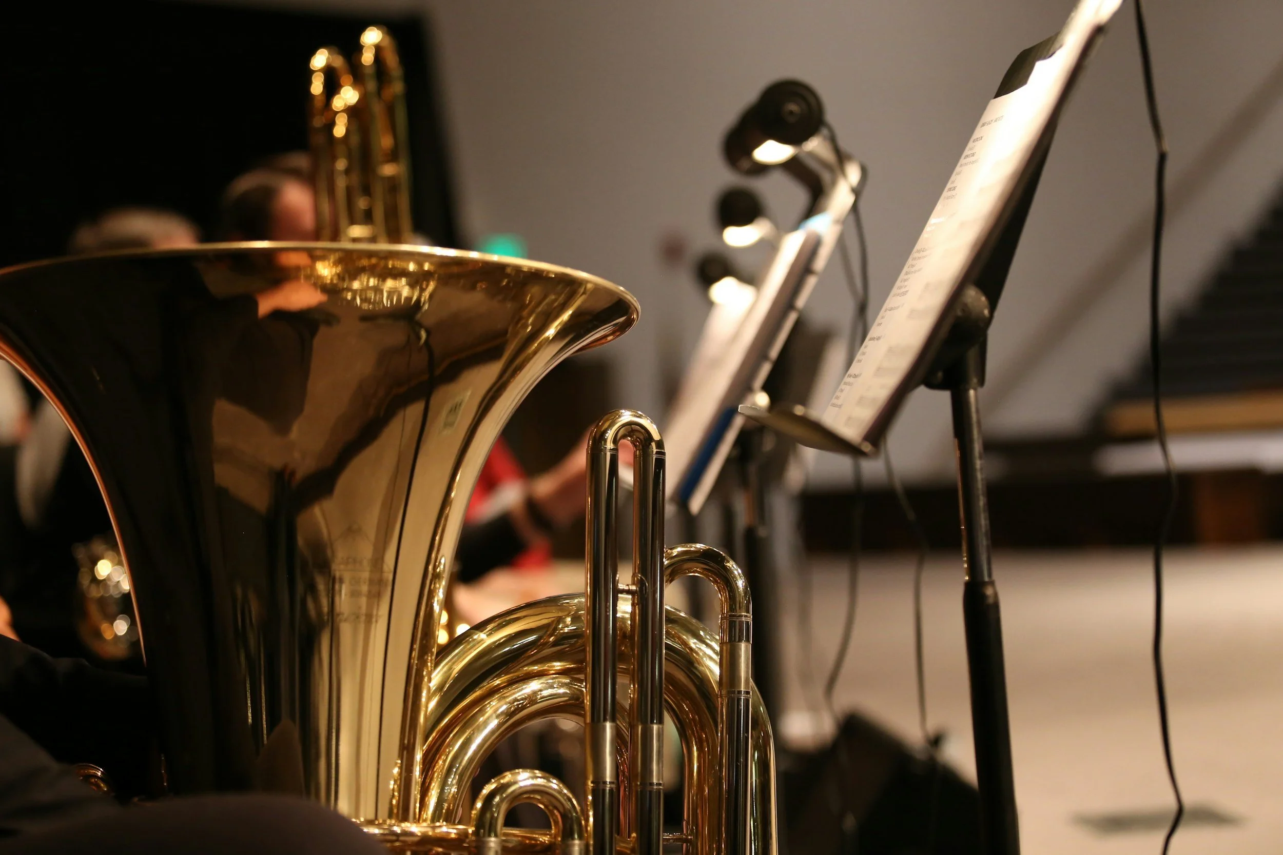 Close-up of a shiny brass tuba in an orchestra setting, with music stands and sheet music in the background.