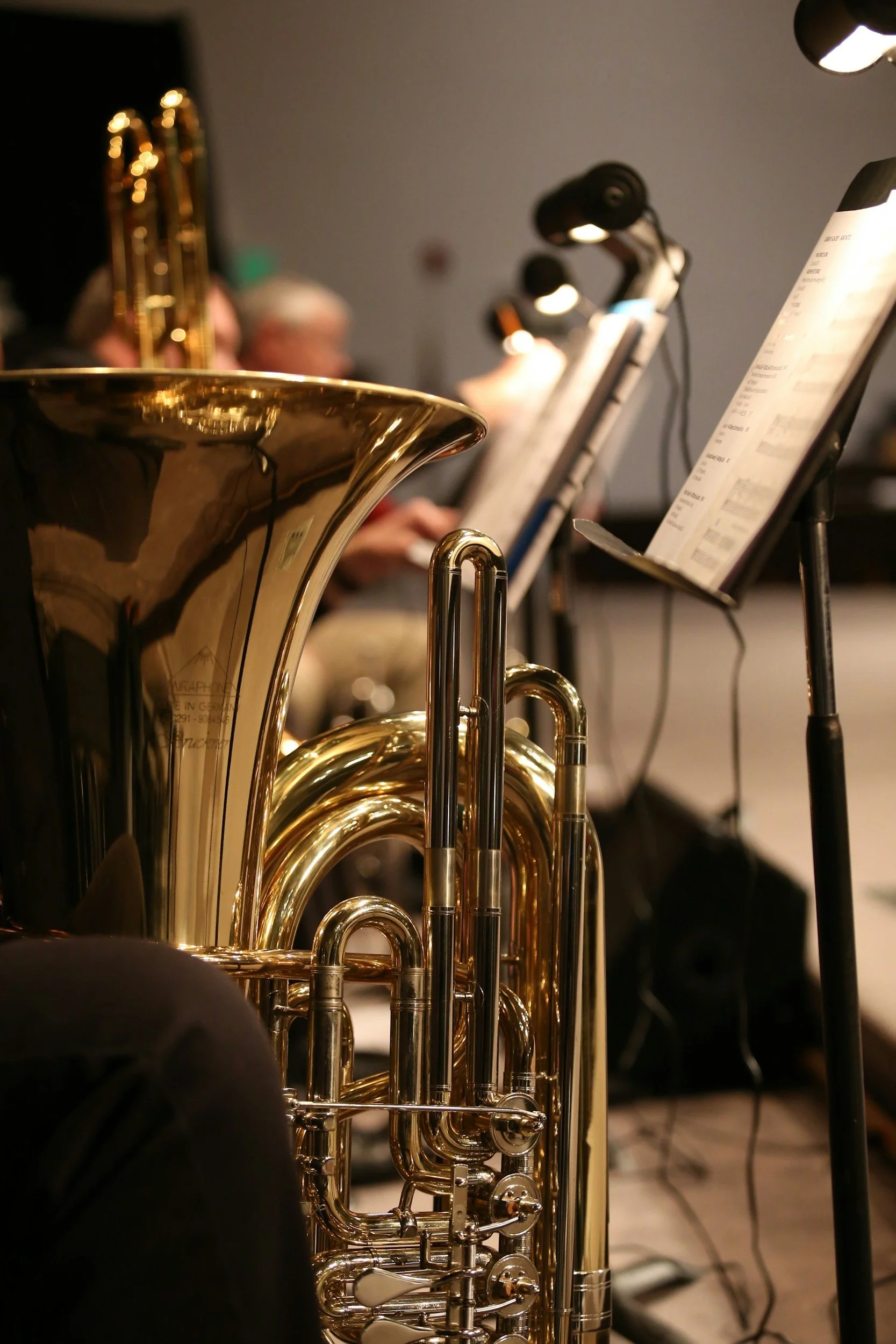 A brass musical instrument, likely a euphonium or tuba, in the foreground with sheet music and music stands in the background during a performance.