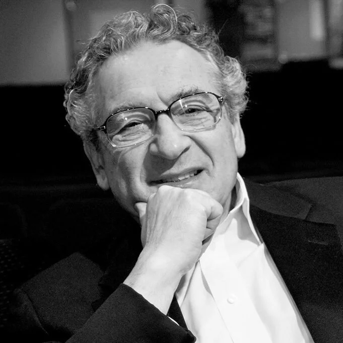 A man with curly hair and glasses smiling with his hand resting on his chin, dressed in a white shirt and dark blazer.