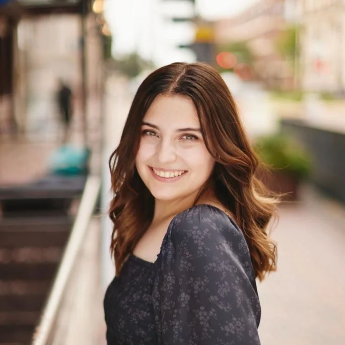 A young woman with long, wavy brown hair and a bright smile standing outdoors on a city street.