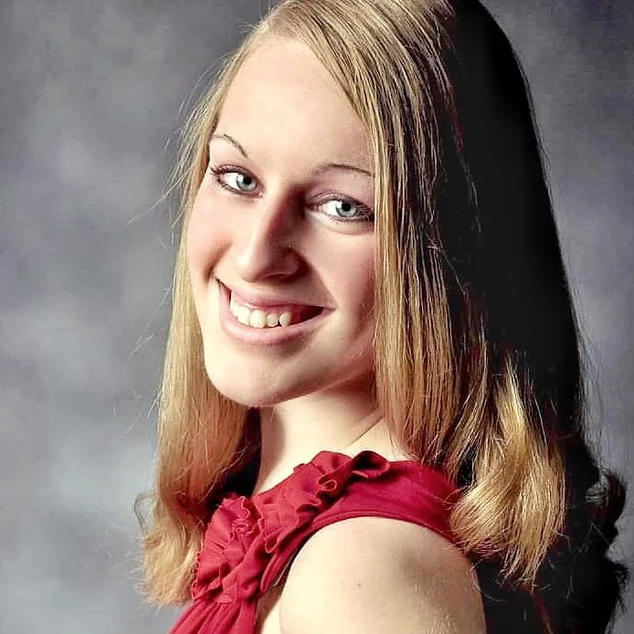Close-up portrait of a young woman with long blonde hair, smiling and wearing a red top, against a gray background.