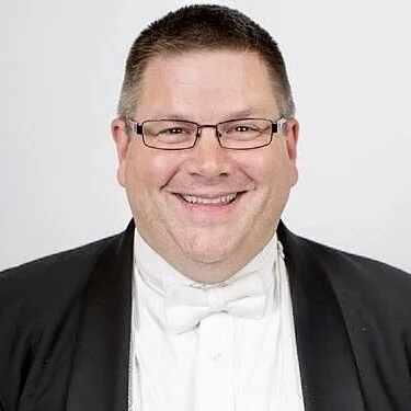 Portrait of a smiling man wearing glasses, a tuxedo, and a white bow tie, with short hair and a light background.